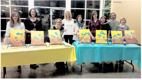 group of nine adults standing in a line and showing their art canvases. They are standing behind two tables, one with a yellow table cloth and the other with a blue table cloth. 