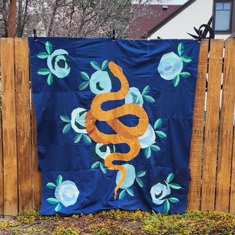 Picture of a snake quilt top attached to a fence. Snake is light golden with a dark golden underbelly, the flowers are green and blue and the background is dark blue.