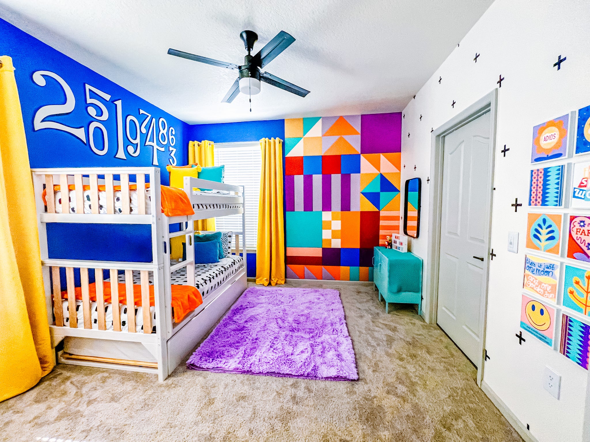 Bedroom with ceiling fan, yellow curtains over two windows, and a purple rug. Geometric Felt Right tiles on back wall, photo collage on right wall, and giant number decals on the left wall.