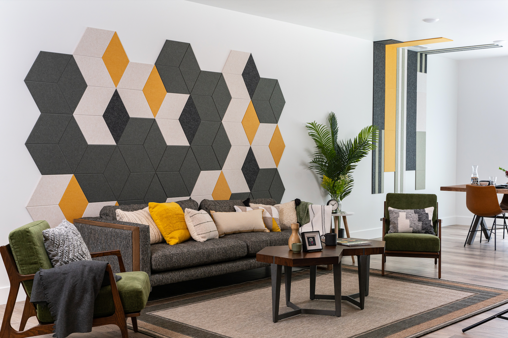 Living room with grey couch and coffee table. Grey, Cream, and Yellow hexagon tiles are on the wall behind the couch. Dining room in the background with Yellow, Cream, and Grey Felt Right tiles on the wall and ceiling in vertical lines.