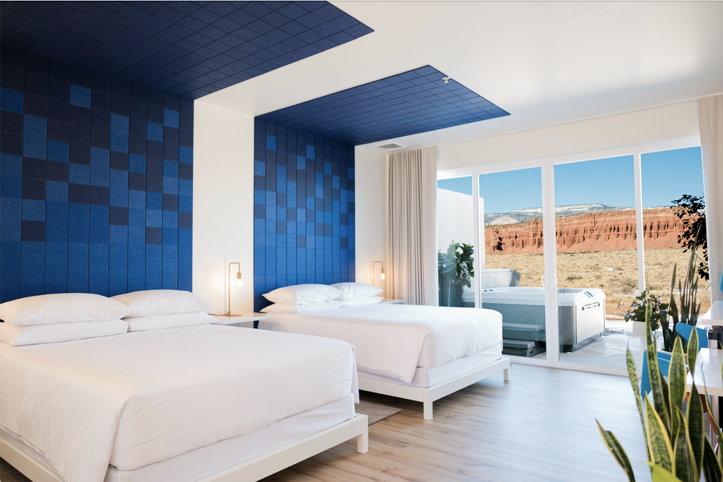 Two beds in a hotel room with a view of red rock out the back glass doors. Blue wall tiles are behind each bed and go onto the ceiling above.