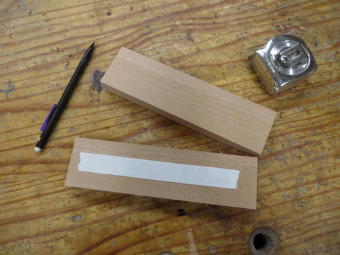 A black mechanical pencil and two pieces of cut wood with a piece of of double sided tape along the length of one of them, and a tape measure all on a wooden workbench