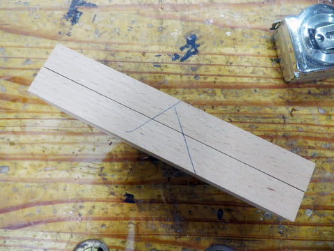 two pieces of cut hardwood stuck together with two perpendicular pencil marks running across them to create a "v" and a measuring tape, all on a wooden workbench