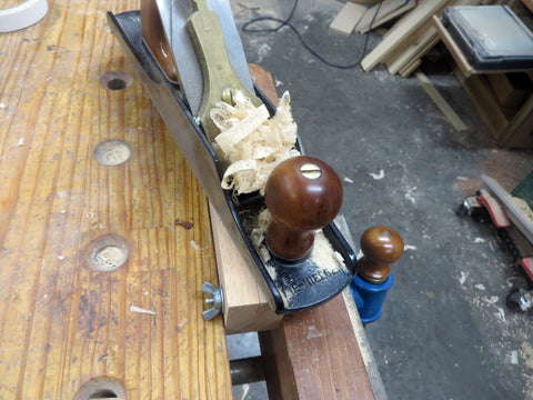A Hand plane on the scraper vise with wood shavings on top of the hand plane. The scraper vise is clamped against the edge of a wooden workbench