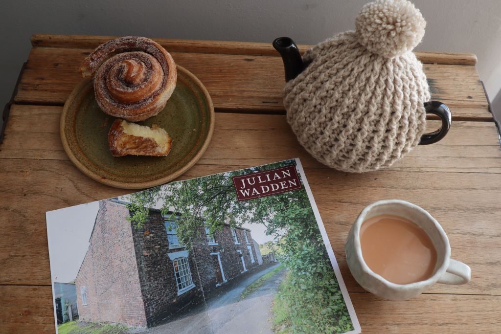 alt="flat lay shot of estate agent particulars, handmade cup and plate, cinnamon roll and brown betty teapot with teacosy. Read The One That Got Away on Bramble and Fox UK hygge shop blog."