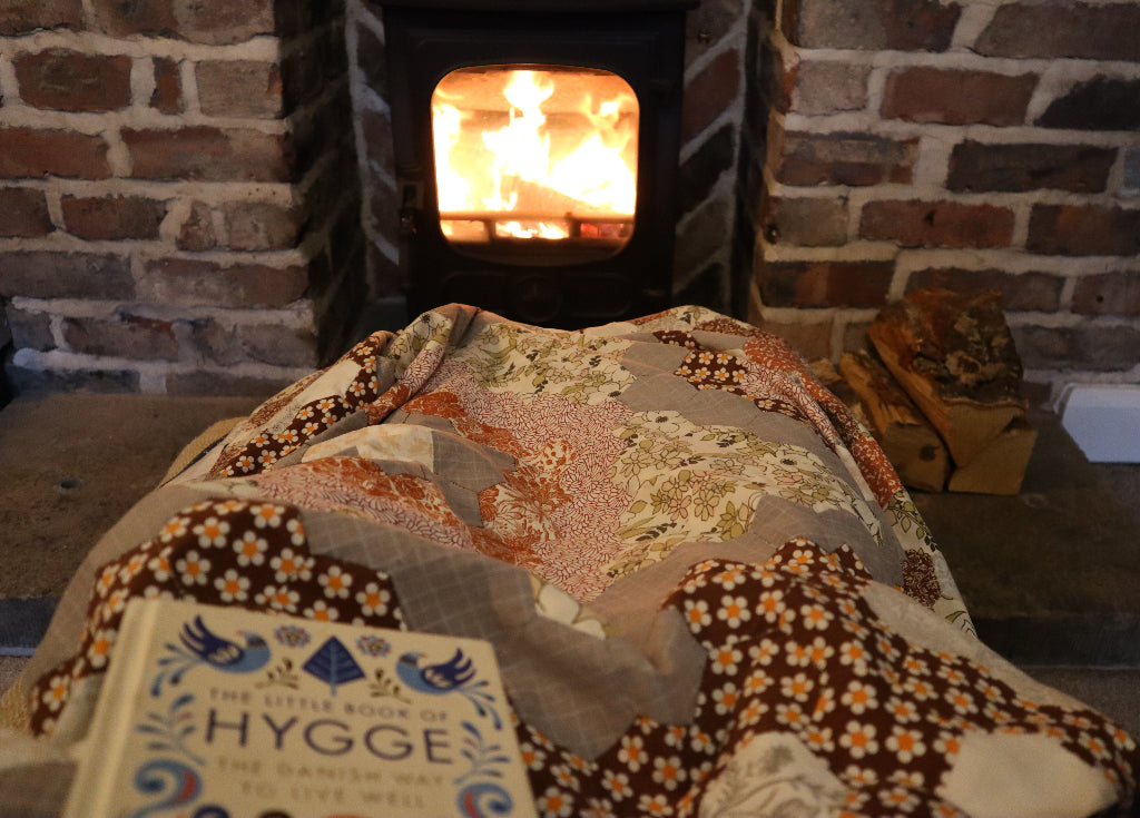 alt="cosy patchwork quilt with wood stove and the little book of hygge by Bramble and Fox UK hygge cottagecore shop"