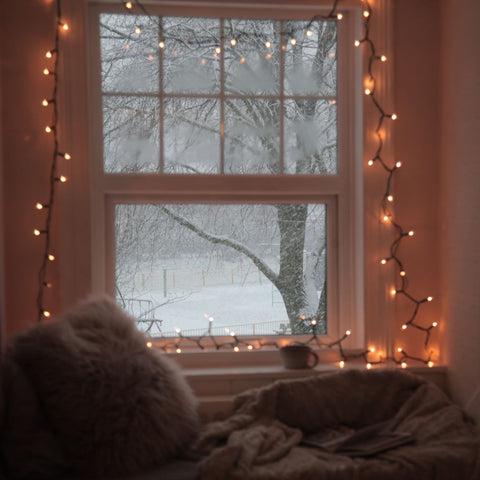 alt="emotional escape rooms and hyggekrogs. A cosy window seat scene with twinkling fairy lights, cushion, blanket and a snowy landscape. Read about 2023's trend for nooks at Bramble and Fox Uk hygge blog."