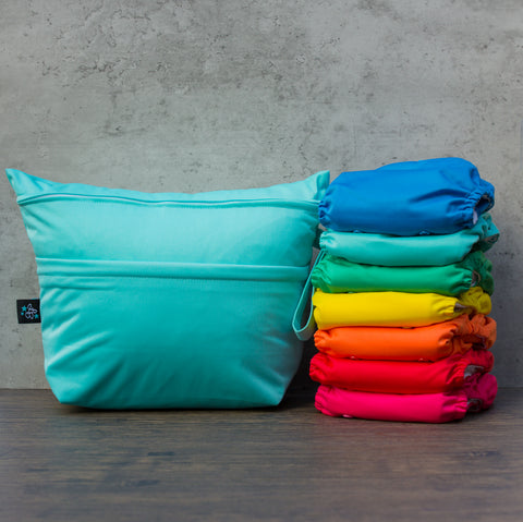 Stack of rainbow one size cloth diapers sits beside an aqua colored Merrily Merrily Quick Trip wet/dry bag