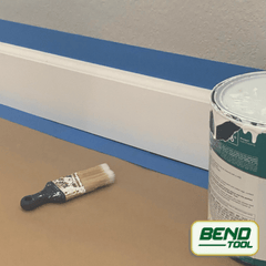 Bend Tool Co. - White profiled baseboard prepped with blue painters tape on hardwood floor, paint and paint brush.