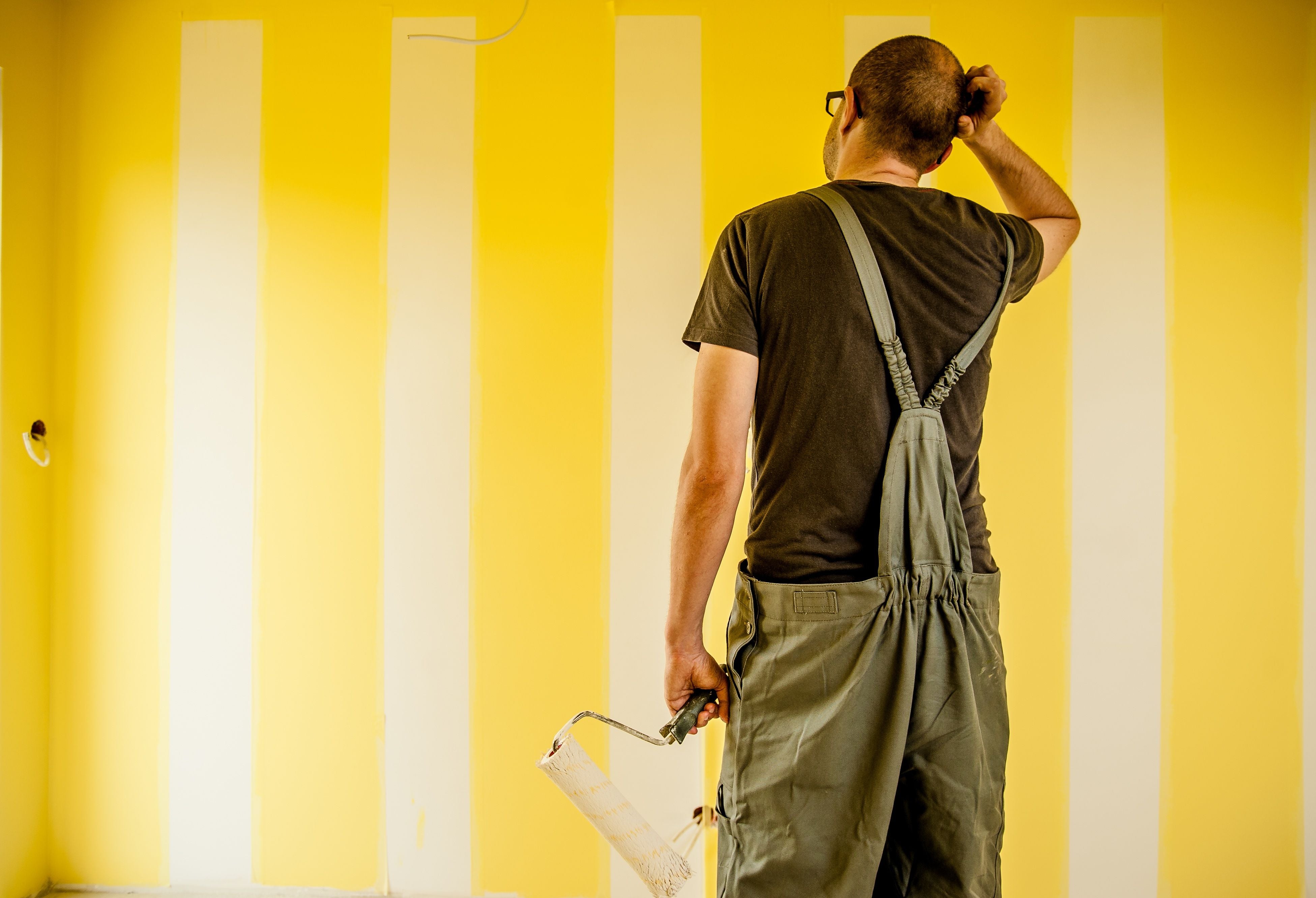 Man stares at the yellow wall where paint is in progress determing to paint baseboards or wall first. Bend Tool Co.