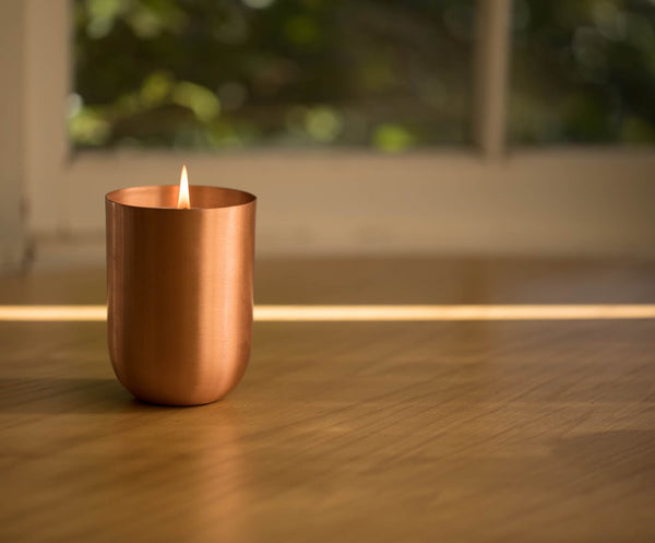large copper jar candle, lit on a pine table with a green leafy background.