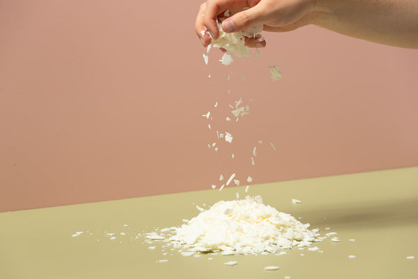 Caucasian hand sprinkling soy wax flakes onto a green table with a pink backdrop