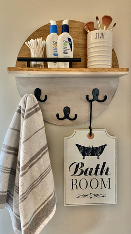 round wall art with shelf holding bathroom lotions and bruches and small green in house shaped pot; hooks hold bath towel and black and white bathroom sign