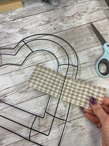 Dark green heart shaped wire wreath form and beige and white gingham ribbon being threaded through the wire