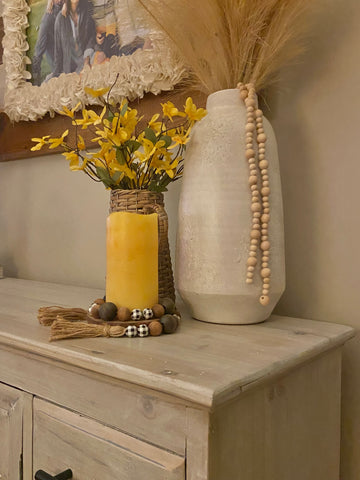 tall white vase with whispy stems sticking out, wicker pitcher with yellow stems and a yellow candle wrapped with black and white beads on top of a white cabinet