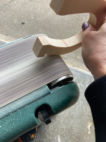 person's hand holding a wooden cut out of a horseshoe on a belt sander as it is sanding the side of the wood