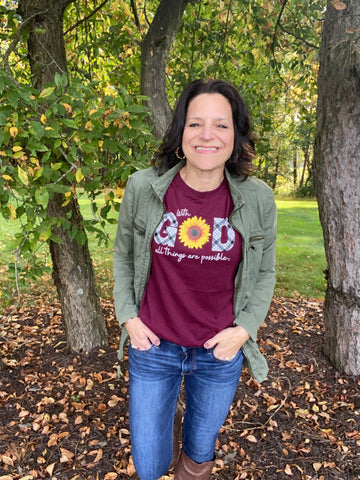 woman wearing jeans and tall boots and army green jacket over a maroon t-shirt that says "With God, All Things are Possible."