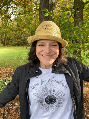 dark haired woman wearing a white t-shirt with black sunflower graphic that says "Shine Like a Sunflower"
