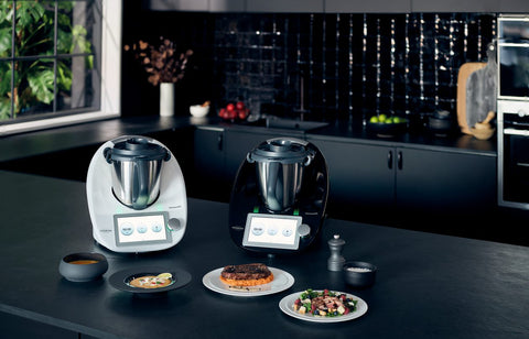 White and black Thermomix machines on kitchen bench with cooked meals surrounding