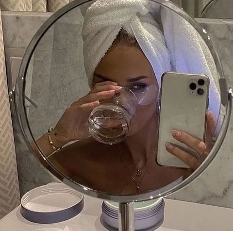 A girl in a wearing a white towel on her head drinking water in front of a mirror