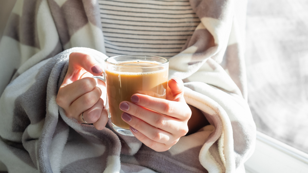 Woman wrapped in white and gray stiped soft blanket holding a cup of coffee with cream between her hands