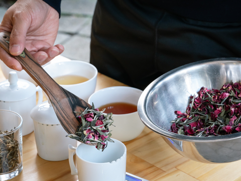Rujani Tea’s Rose Green blend is being tasted along with other artisanal teas. A spoonful of Rose Green Tea is being poured into a tasting cup.