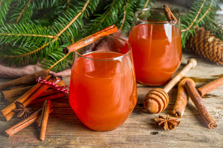 two classes with holly jolly tea mocktail in them stand on a table surrounded by cinnamon, honey, and tree boughs