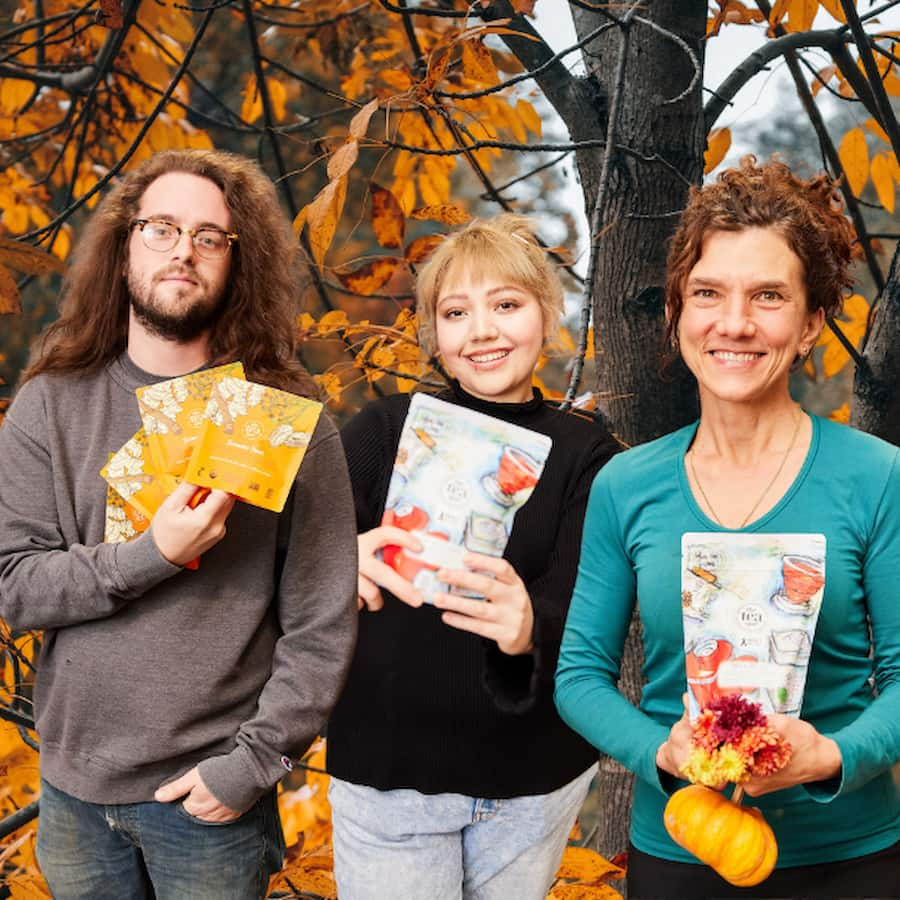 three people stand holding tea bags in front of fall leaves