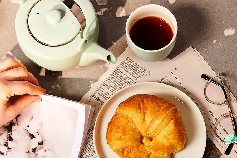 a croissant, newspaper, tea pot and tea cup sit on a table