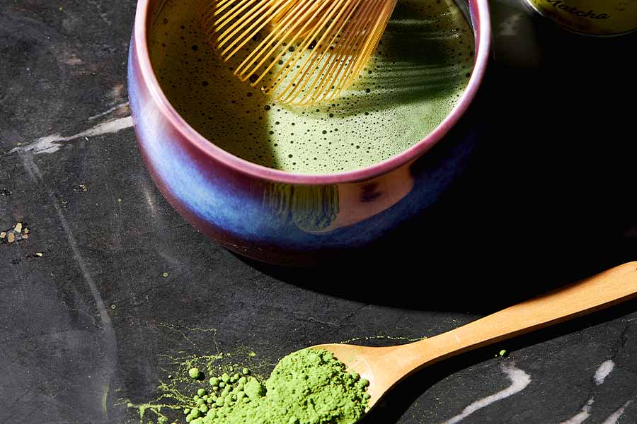 a bamboo whisk whisks matcha in a bowl with a spoon with matcha green tea powder on it to the side