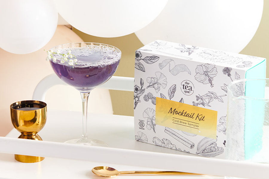 a purple tea mocktail sits beside a box reading mocktail kit with balloons behind it
