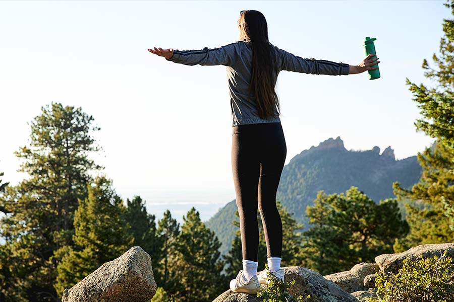 a girl stands on a mountain with her hands out holding a tea tumbler