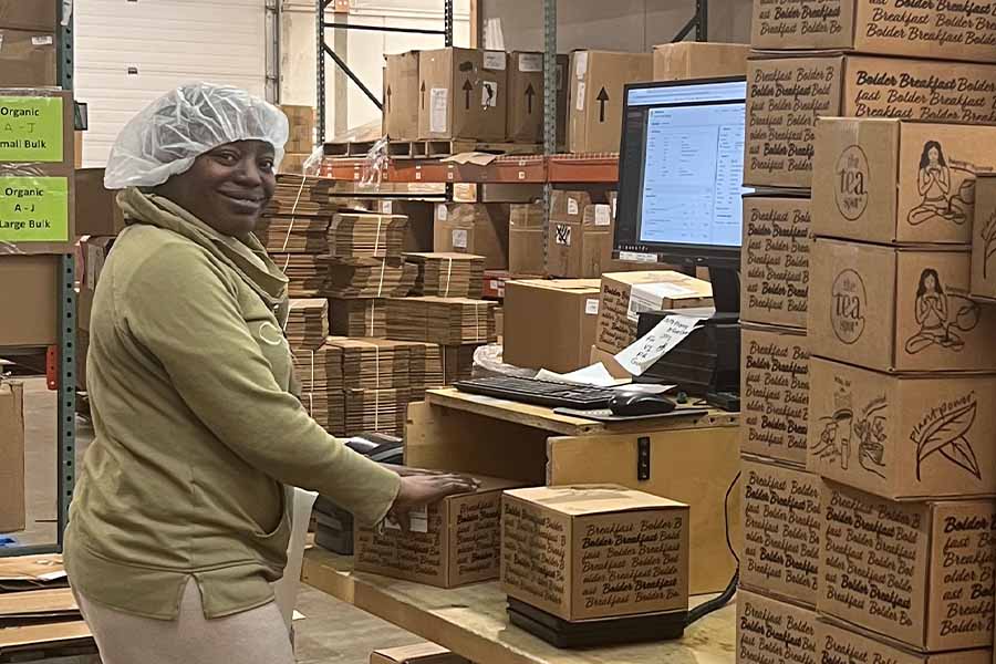 a woman stands at a computer with a hair net on with shipping boxes around her