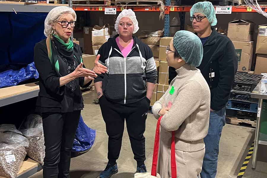 a team of people stand with hairnets in a warehouse listening to a woman talk