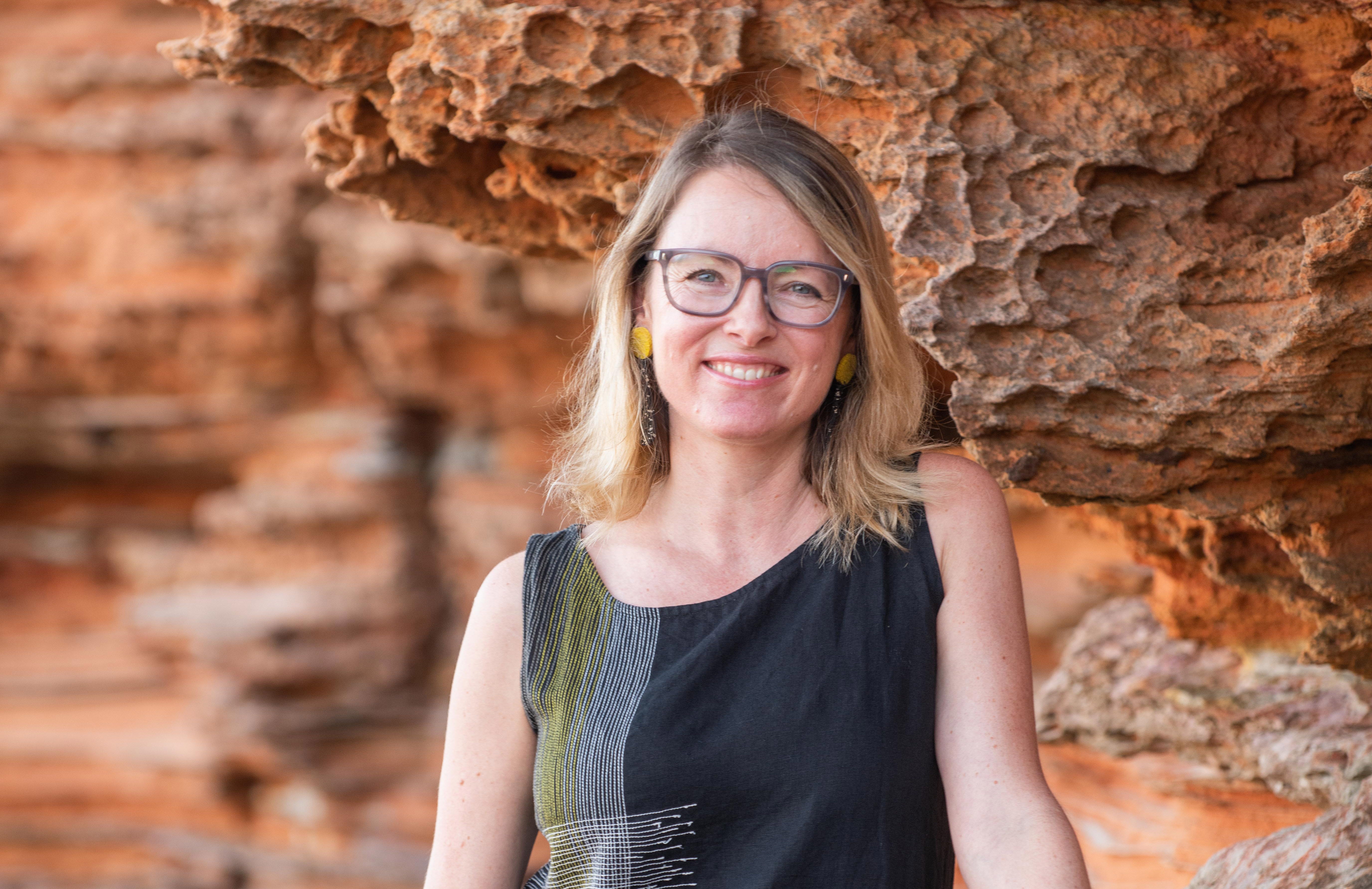 Anna is a thin white woman with blond hair. She is standing in front of limestone rocks. She is wearing glasses and smiling at the camera.