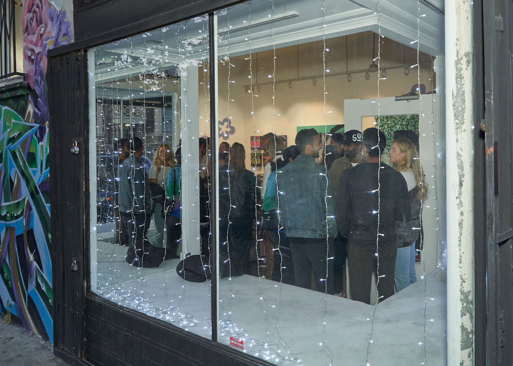 Photograph of people captured through a glass wall with Christmas lights during Ernie Steiner's "Figuratively Speaking" solo exhibition Opening Reception at Voss Gallery, San Francisco, April 22, 2022.