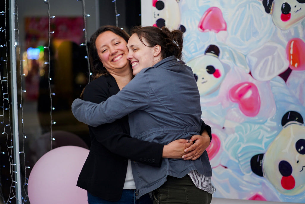 Warm embrace between two friends sharing a joyful moment at a San Francisco art gallery event, with whimsical contemporary art in the background, promoting local artists at Voss Gallery.