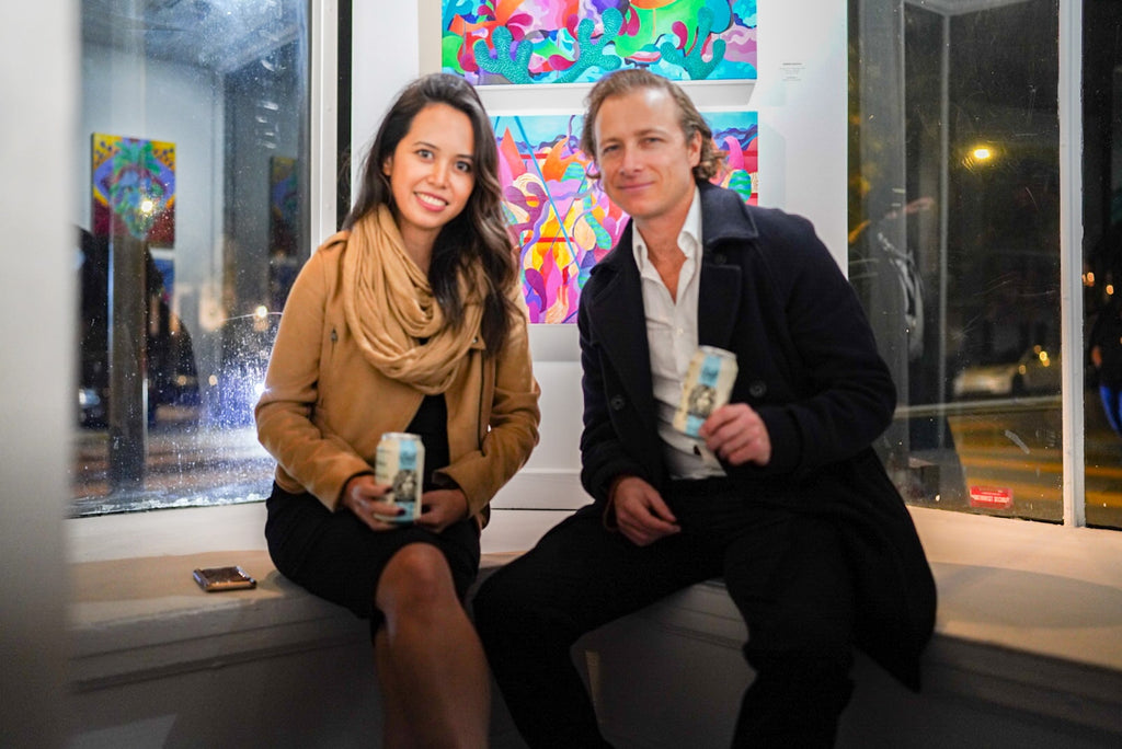 Photograph of two people holding canned drinks during Jennifer Banzaca & Joshua Nissen King's "Mirage" duo exhibition Opening Reception at Voss Gallery, October 22, 2021.