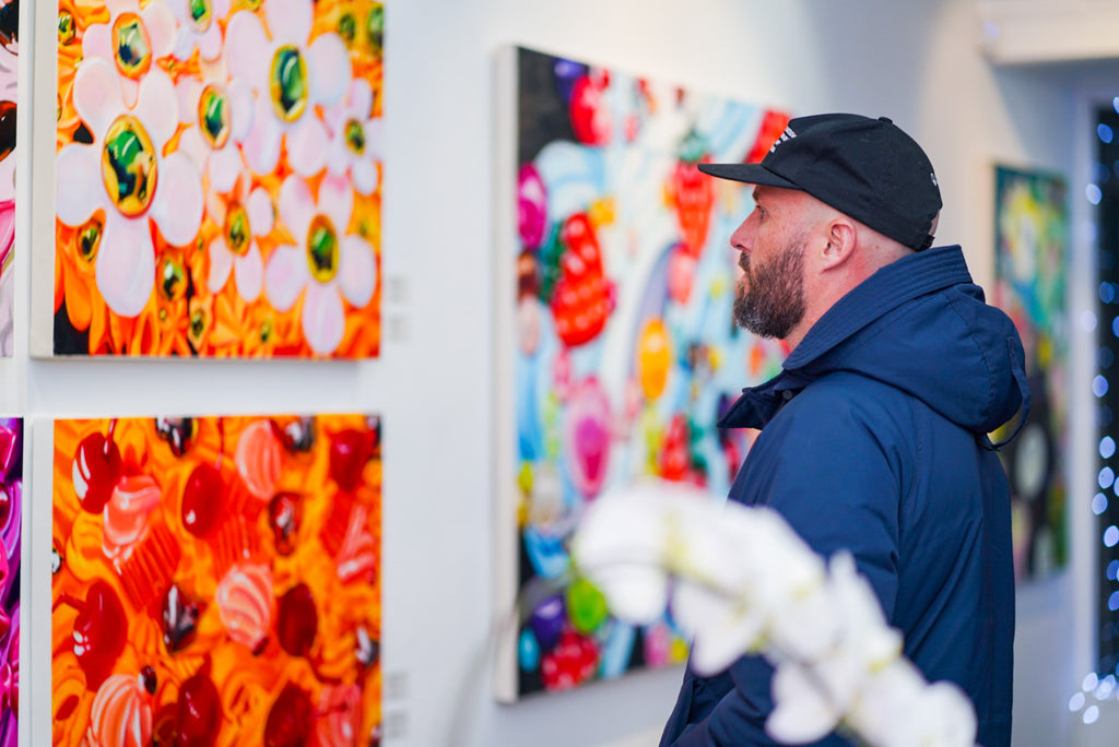 Man in a dark jacket observing colorful abstract paintings in an art gallery, showcasing vibrant floral and balloon-like shapes on canvas, indicative of the lively art scene at Voss Gallery, San Francisco.
