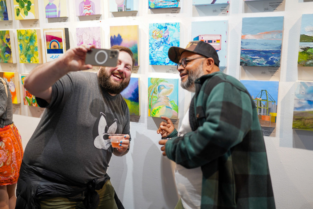 Photograph of two men taking a selfie with paintings during the Opening Reception of the Postcards from Paradise Juried Group Exhibition at Voss Gallery in San Francisco, May 26, 2023.