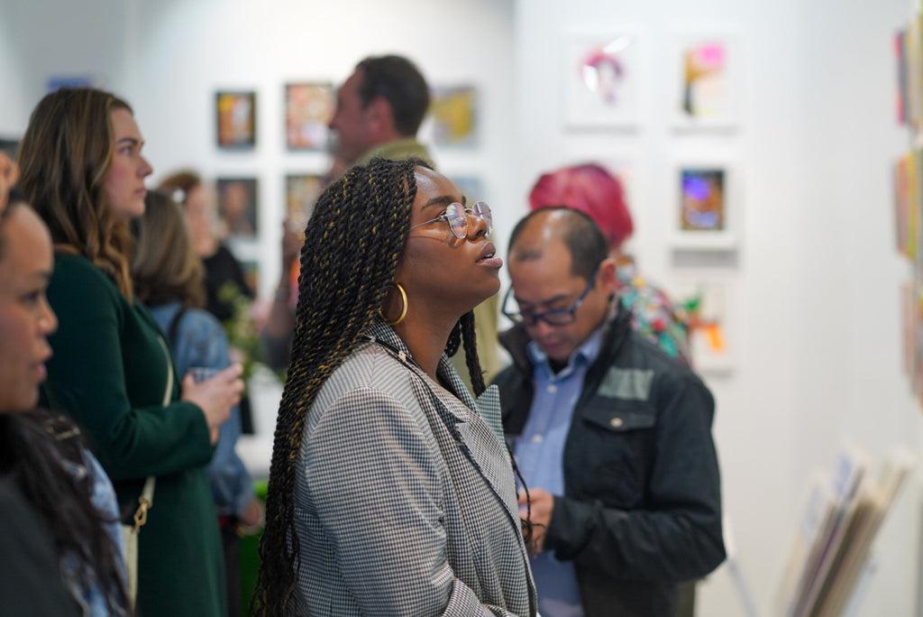 Photograph of a lady and some guests viewing artwork during the Opening Reception of the Postcards from Paradise Juried Group Exhibition at Voss Gallery in San Francisco, May 26, 2023.