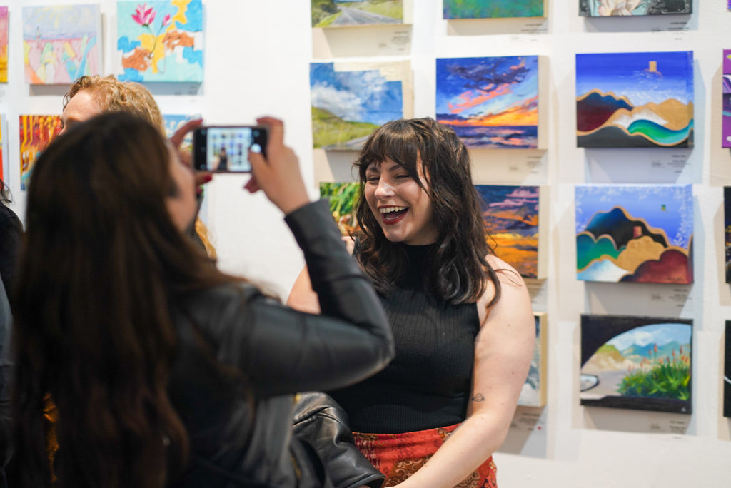 Photograph of a woman being photographed by another woman with her phone during the Opening Reception of the Postcards from Paradise Juried Group Exhibition at Voss Gallery in San Francisco, May 26, 2023.