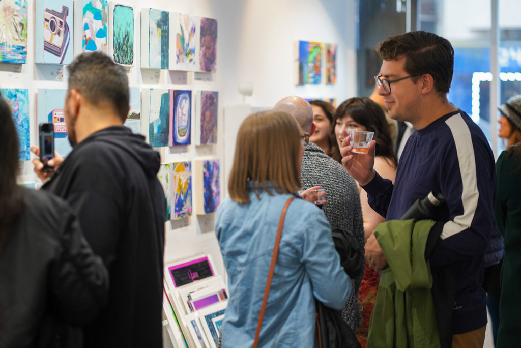 Photograph of people checking out hanged paintings during the Postcards from Paradise Juried Group Exhibition Opening Reception at Voss Gallery in San Francisco, May 26, 2023.