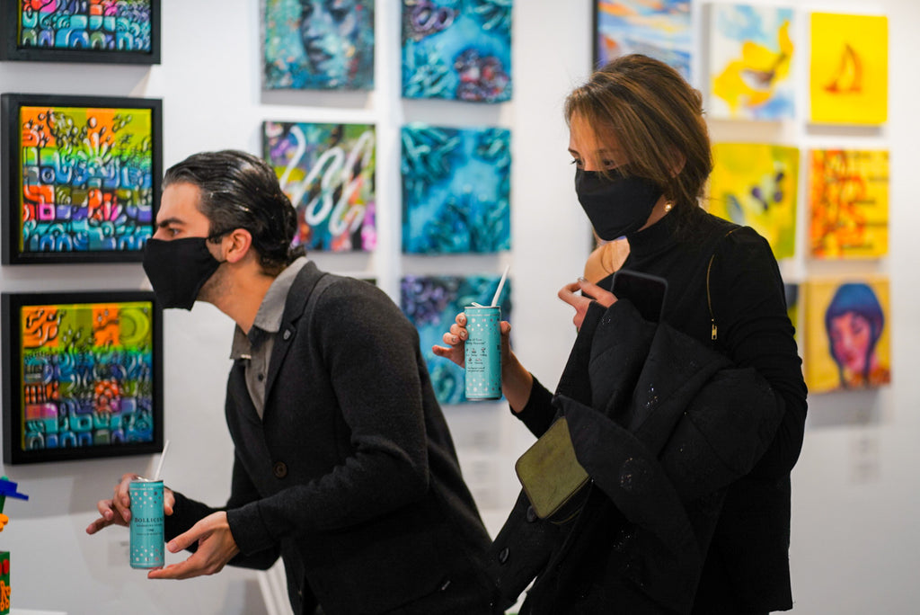 Photograph of two people holding canned drinks and viewing artwork during the "It's BASL, Baby!" group exhibition VIP Young Collectors Event at Voss Gallery, San Francisco, December 4, 2020.