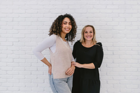 Two women of different body types, one in layered shirts, in front of a white brick wall