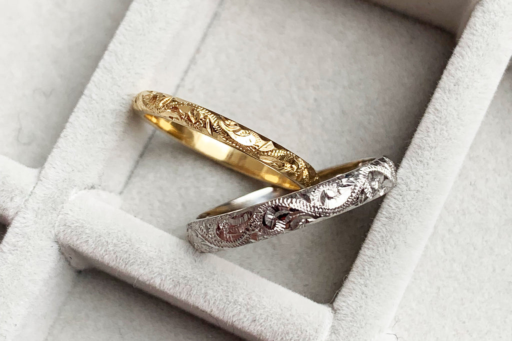 A pair of slender court-shaped wedding bands hand engraved with scrolls, resting against one another jauntily. The top band is recycled yellow gold and the bottom is recycled platinum