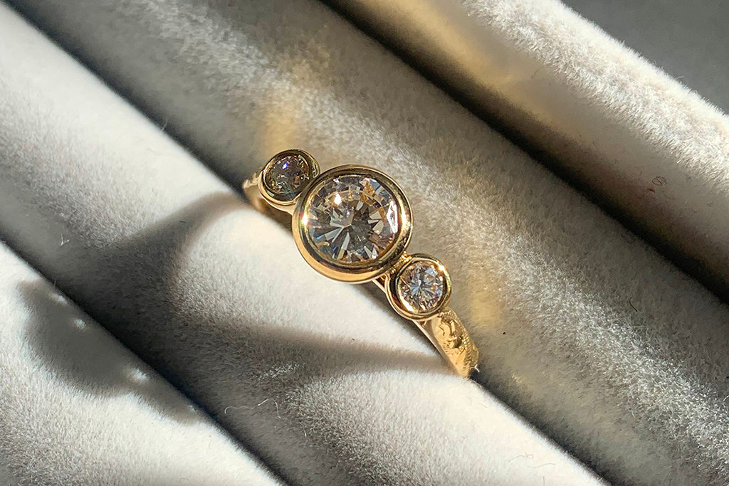 An ethical bespoke engagement ring, cast in recycled gold and set with a trilogy of our client's own reclaimed vintage old cut diamonds, secured in bezel settings and a hand engraved band