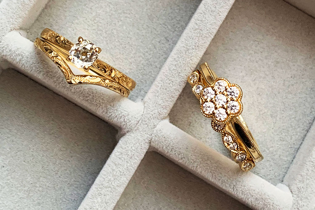 A recycled yellow gold and old cut diamond solitaire Athena engagement ring paired with a scroll-engraved Wishbone wedding band. Alongside it, a vintage-inspired Daisy cluster engagement ring with a slender marquise diamond-set Amare wedding band
