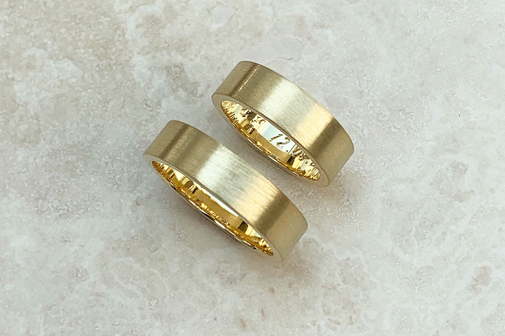 His and hers - two wide, flat-profile wedding bands in yellow Fairtrade Gold, finished with a matte texture and hand engraved with private messages on their inner bands