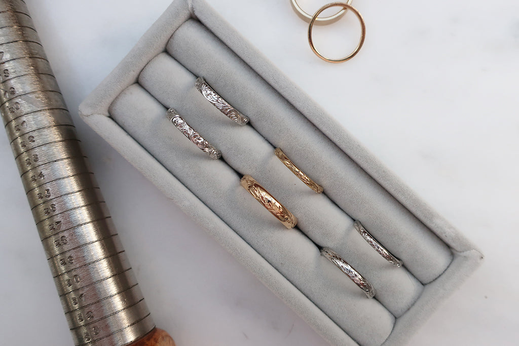 A selection of hand engraved ethical gold and platinum wedding bands lined up in a grey ring cushion